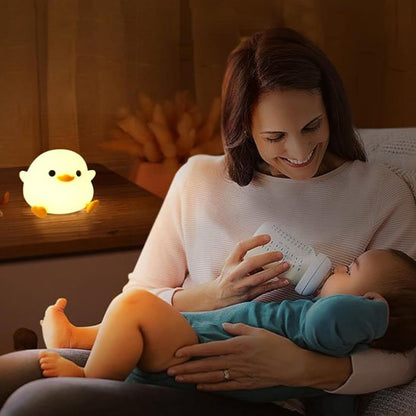 Sur la table de chevet, la veilleuse bébé en forme de poussin éclaire doucement pendant que la mère observe son bébé calme en buvant son biberon.